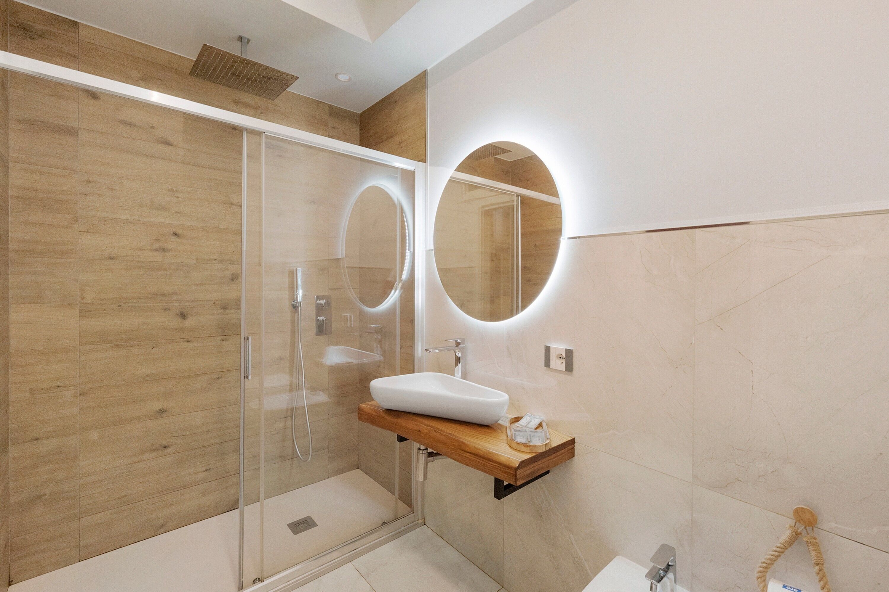 A modern bathroom with a glass-enclosed shower, a wooden vanity with a white sink, and a round mirror with integrated lighting.