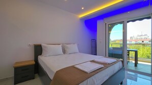 A modern bedroom with a large bed, a nightstand, a wooden bench, and a view of the outdoors through a sliding glass door. - 15 - Alexandros Apartments by SDR (Paralia)