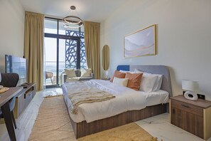 A modern hotel room with a large bed, a desk, a chair, a TV, and a view of a cityscape through a large window. - Bloomfields Studio - The Gate Masdar (Abu Dhabi)