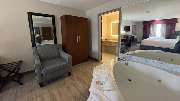 A hotel room with a large bed, a sitting area with a chair, a wooden wardrobe, a bathroom with a bathtub, and a mirror.