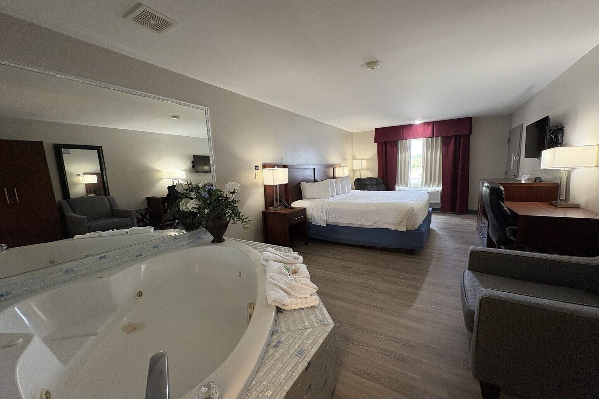 A hotel room with a large bathtub, a bed with a blue headboard, a desk with a chair, and a mirror on the wall.