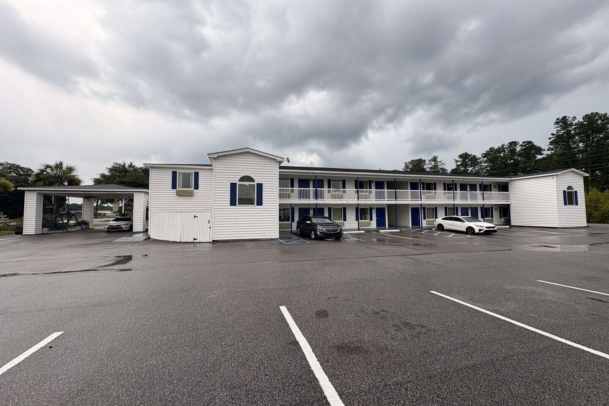 Un motel composé d’un bâtiment principal et de deux annexes plus petites, d’un espace de stationnement couvert et d’un grand parking extérieur.