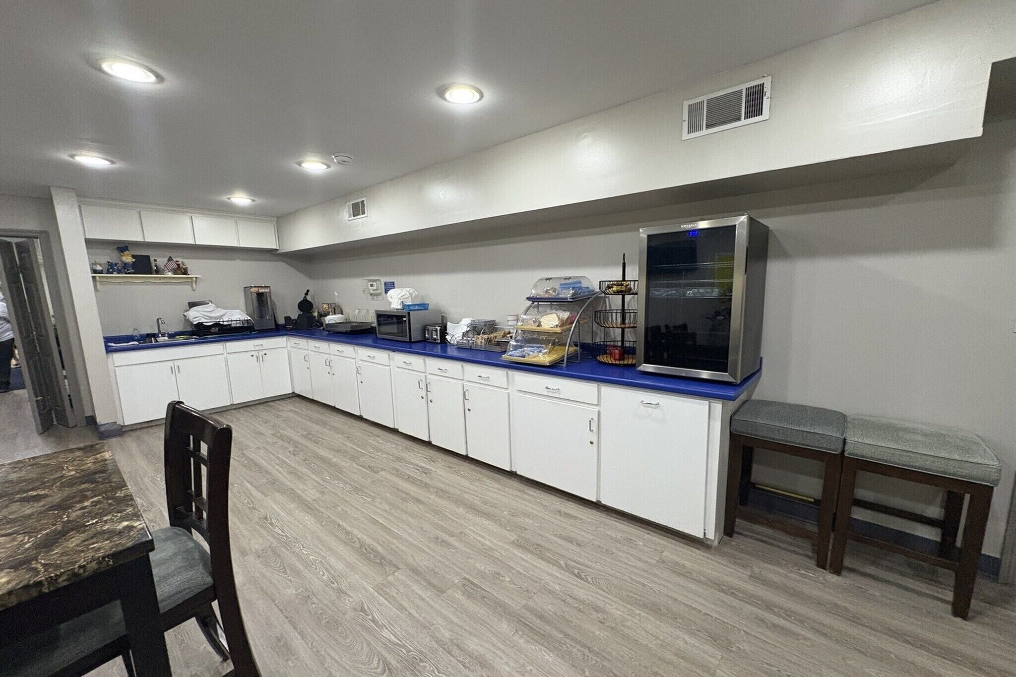 Una cocina con armarios blancos, encimera azul, refrigerador y comedor con mesa y sillas.