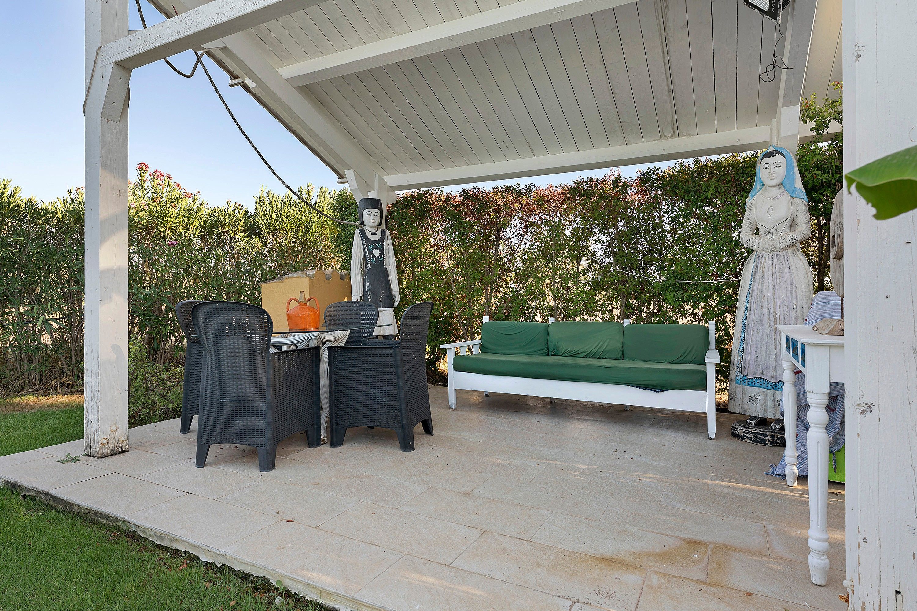 Un patio exterior con una pérgola blanca, un sofá verde, sillones de mimbre y una mesa con un florero.