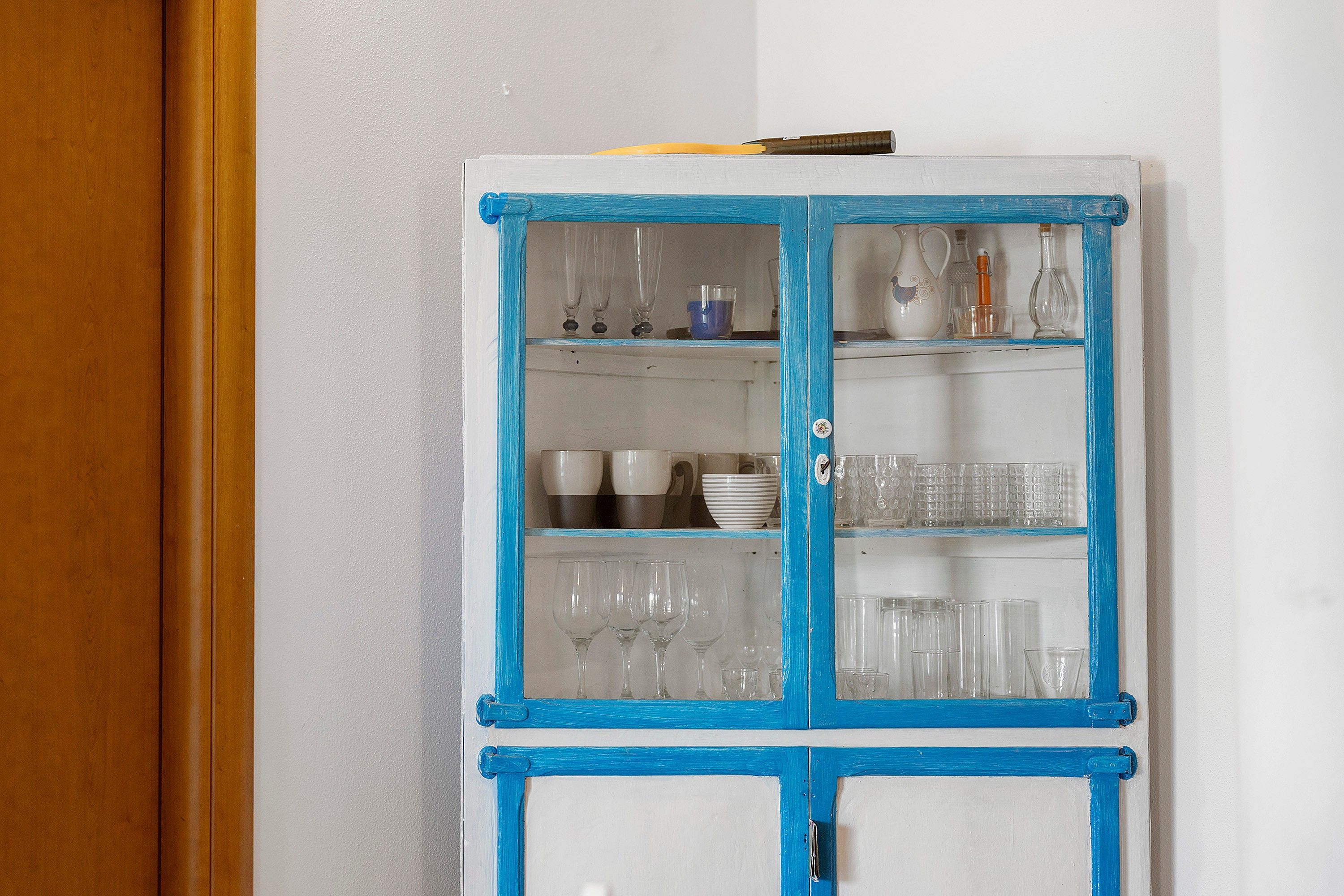 Un meuble blanc avec des bordures bleues et des portes en verre, contenant divers objets en verre et de la vaisselle.