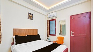 A hotel room with a bed, a red door, an air conditioning unit, a window with curtains, and a small table with a chair. - Classic Agp Home Stay (Chennai)