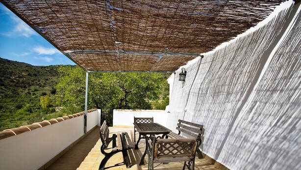 Un patio techado con una mesa y bancos, vista a colinas verdes y una pared blanca.