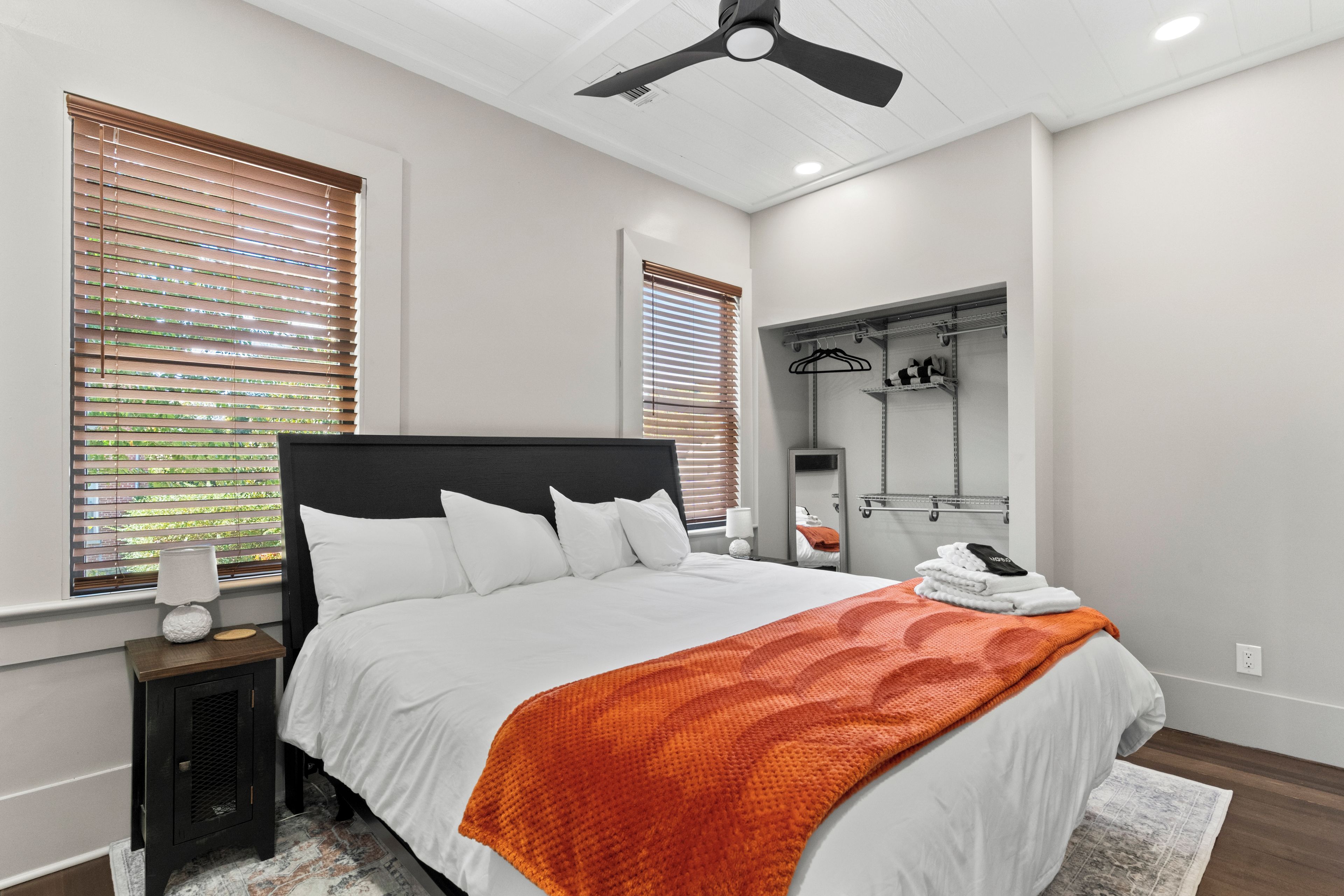A bedroom with a bed, a nightstand, a lamp, a ceiling fan, and a walk-in closet with hanging clothes.