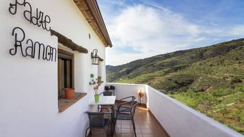 A balcony with a table and chairs, a potted plant, and a view of a green valley.