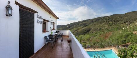 A balcony with a pool, a table, chairs, and a view of a hilly landscape.