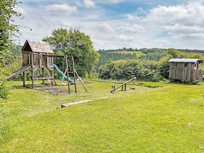 Une aire de jeux avec un toboggan et des balançoires, une structure en bois et un petit abri dans un champ herbeux.
