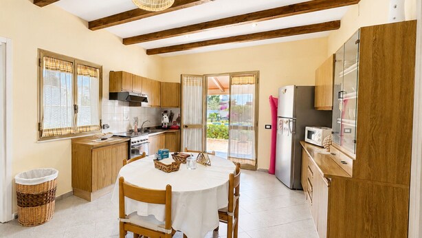 Una cocina con vigas de madera, una mesa de comedor con sillas, un refrigerador y un ventanal con cortinas.