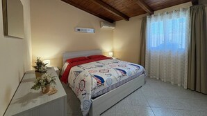 Un dormitorio con techo de madera, una cama con colcha de temática náutica, una mesita de noche con lámpara y una ventana con cortinas.