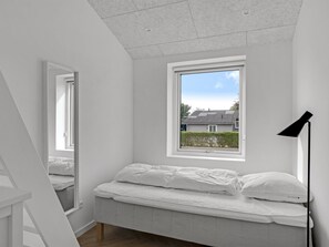 Quarto minimalista com uma cama de solteiro, janela com vista para uma casa e um abajur de chão preto.