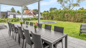 Un área de comedor al aire libre con techo, una mesa larga y sillas, rodeada por un césped cuidado y un seto.