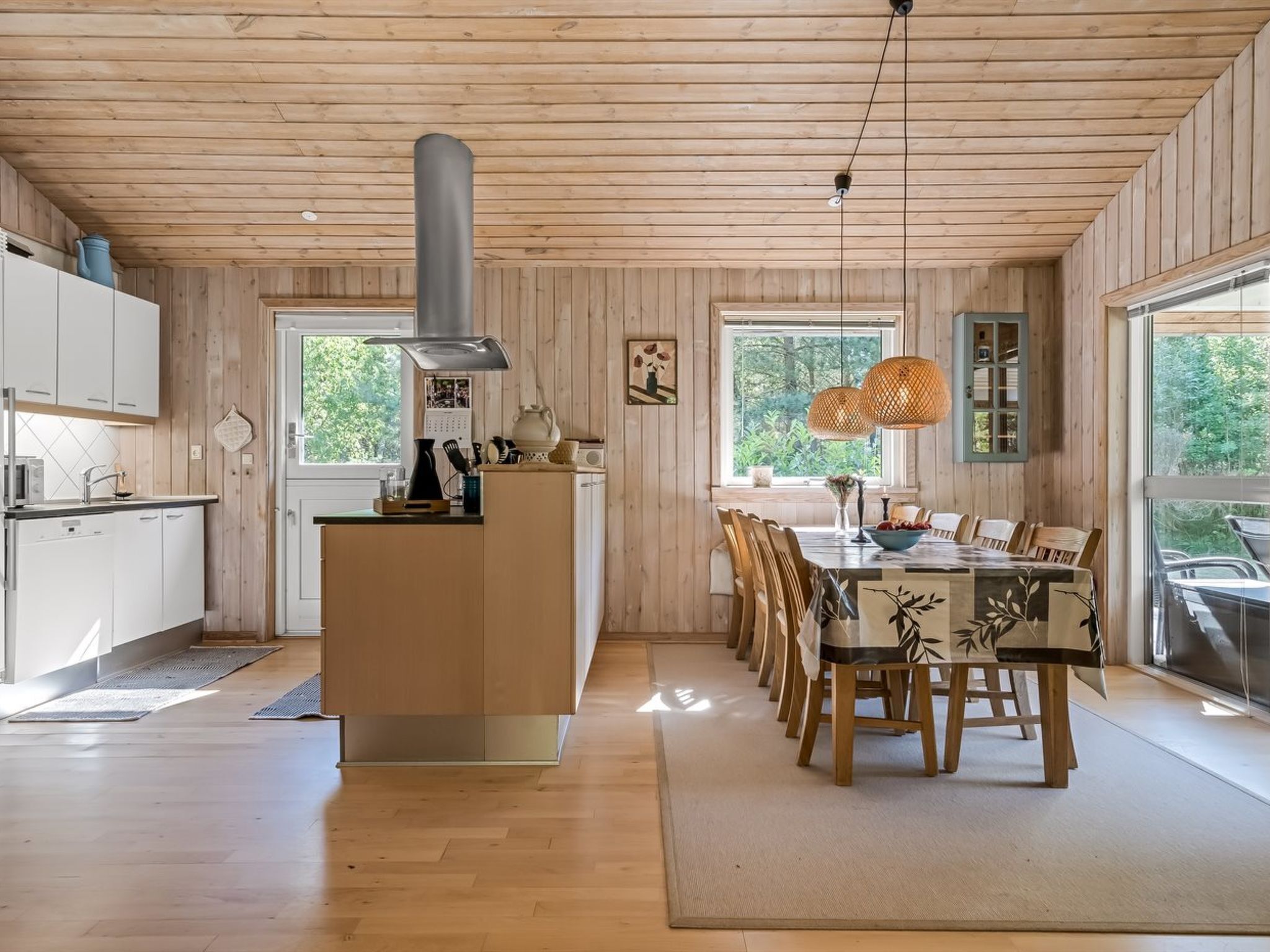 A wooden cabin interior with a kitchen, dining area, and large windows.