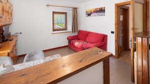A cozy living room with a wooden table, a red sofa, a window with curtains, and a door leading to another room. - Apartment 'Michela' with Mountain View and Balcony (Sant'Anna)