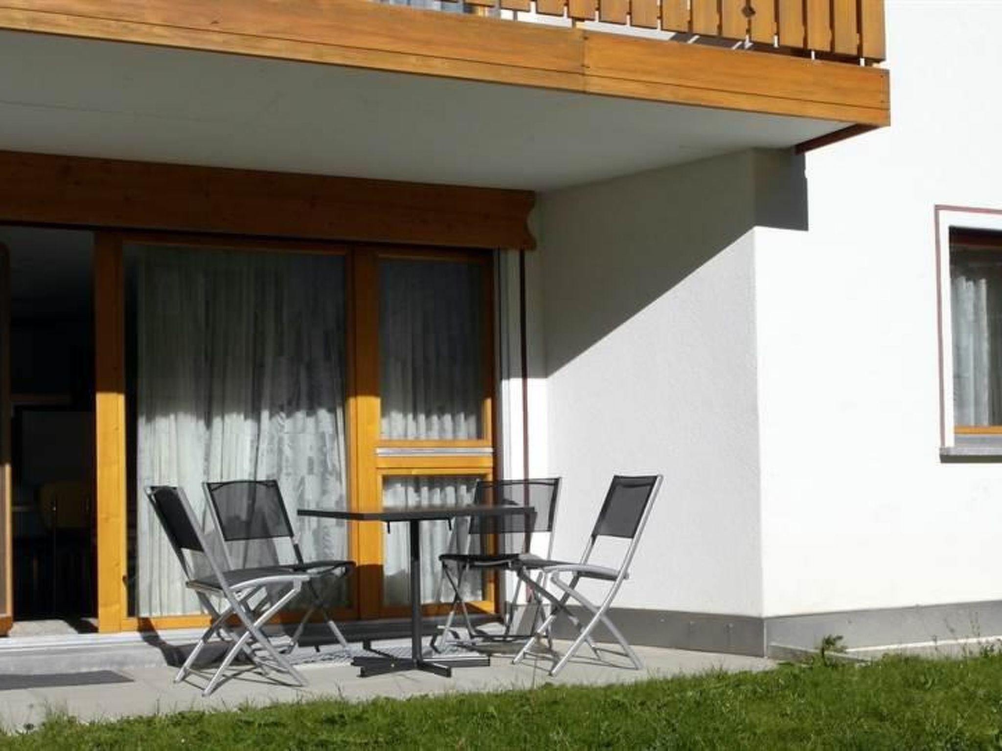 Un área de estar moderna al aire libre con una mesa y cuatro sillas, un balcón de madera y una puerta corrediza de vidrio que da a una habitación.