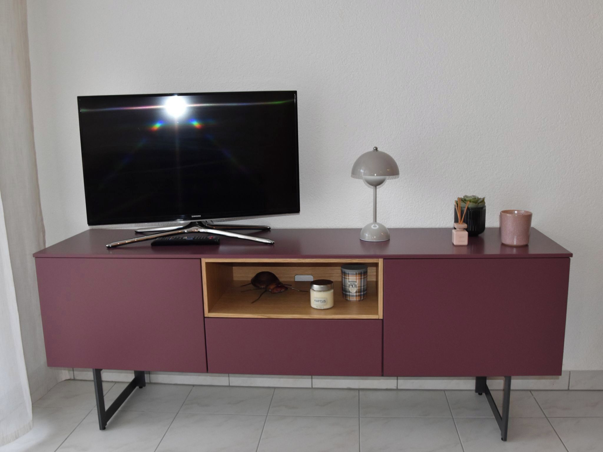 Un téléviseur à écran plat fixé au mur, au-dessus d’un meuble de couleur bordeaux, avec une étagère en dessous.