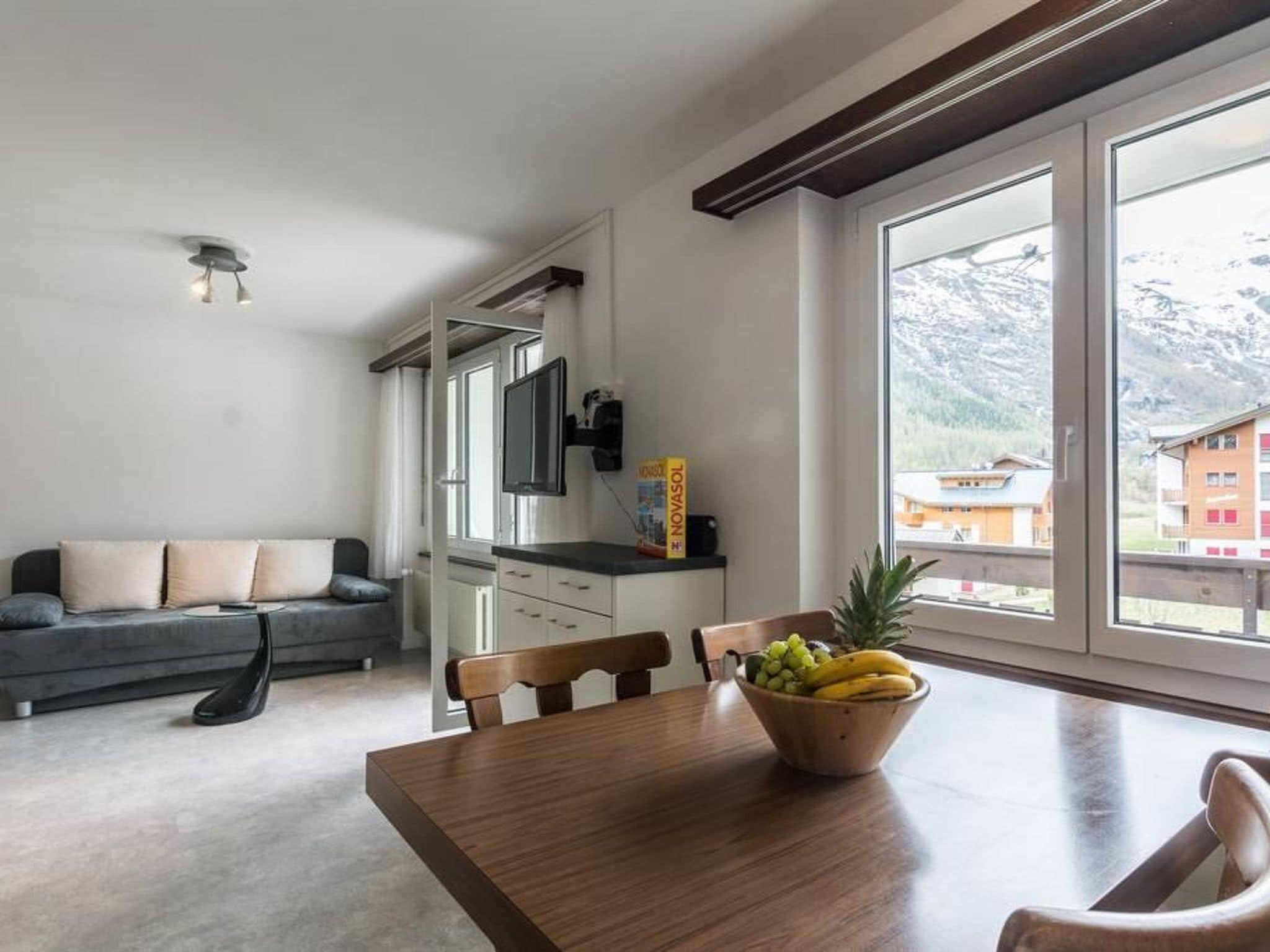 A modern living room with a grey sofa, a wooden dining table with a fruit bowl, and a kitchen area with a flat-screen TV.
