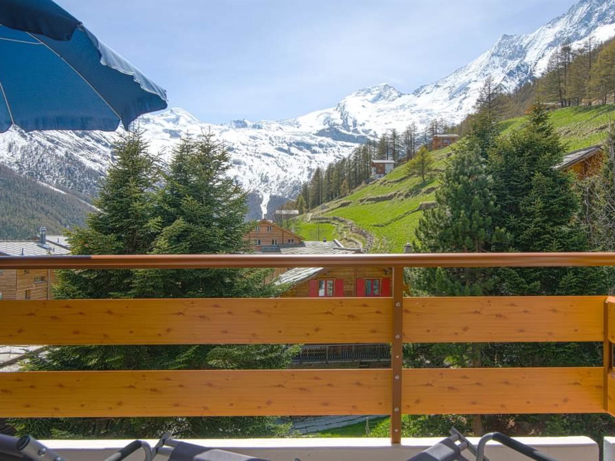 A wooden balcony with a view of a mountainous landscape, including snow-capped peaks and a valley with buildings.