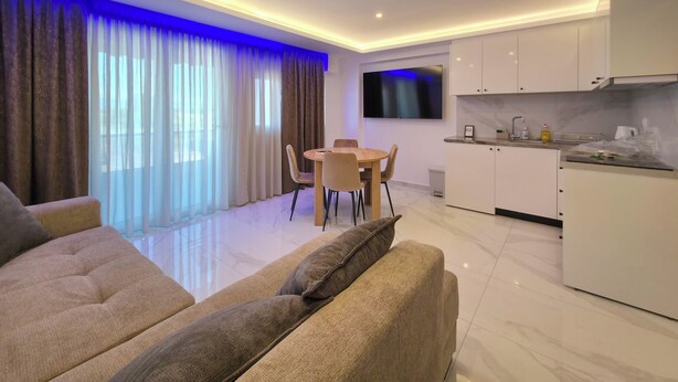 Ein modernes Wohnzimmer mit Sofa, einem Esstisch mit Stühlen, einer Küchenzeile mit Schränken und einem an der Wand befestigten Fernseher.