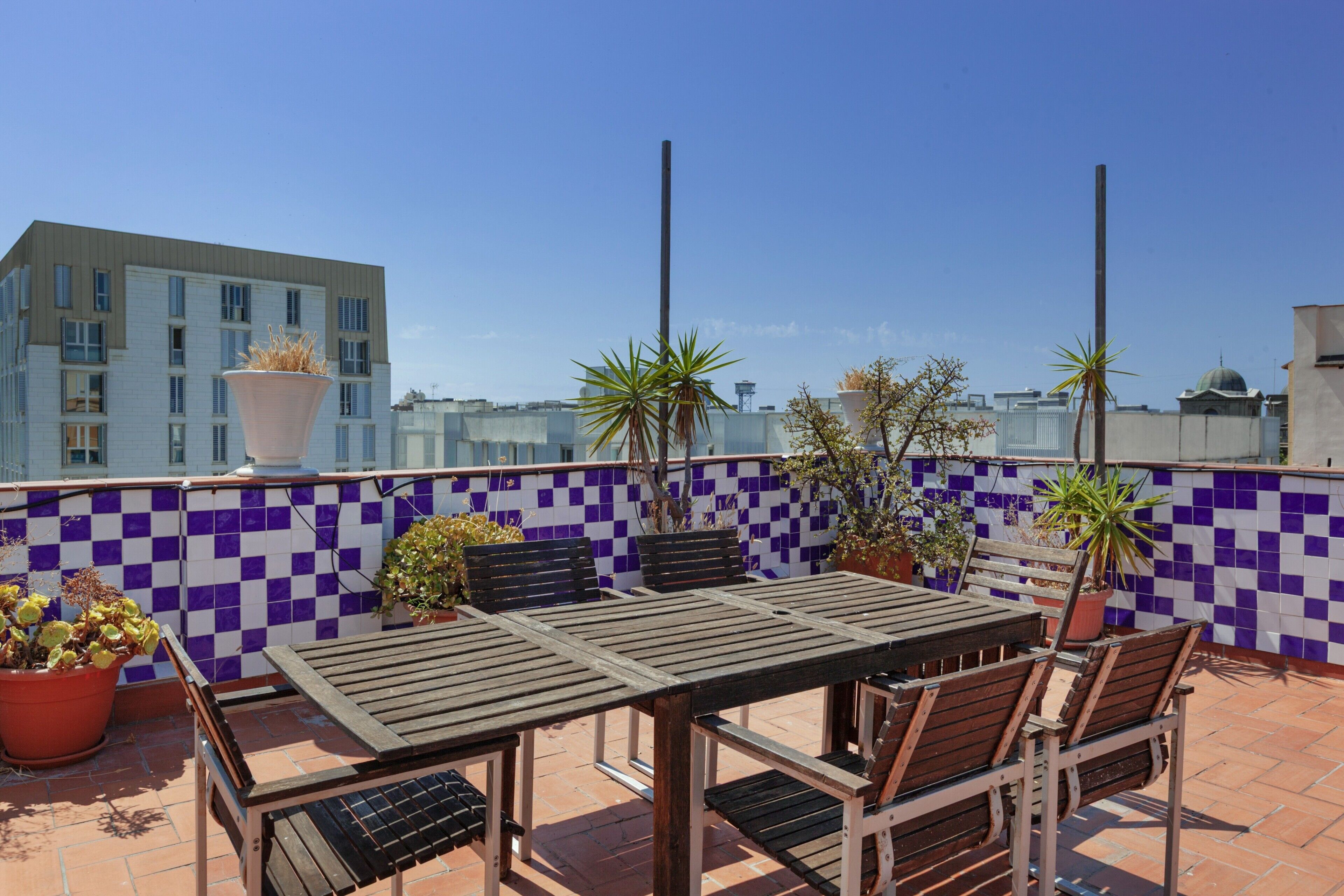 Eine Dachterrasse mit einer Wand aus Schachbrettfliesen, einem Holztisch mit Stühlen, Topfpflanzen und Blick auf eine Stadtansicht.