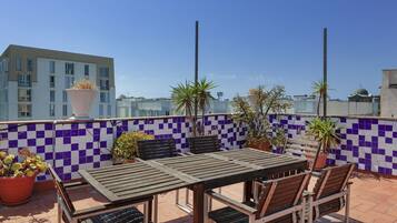 Eine Dachterrasse mit einer Wand aus Schachbrettfliesen, einem Holztisch mit Stühlen, Topfpflanzen und Blick auf eine Stadtansicht.