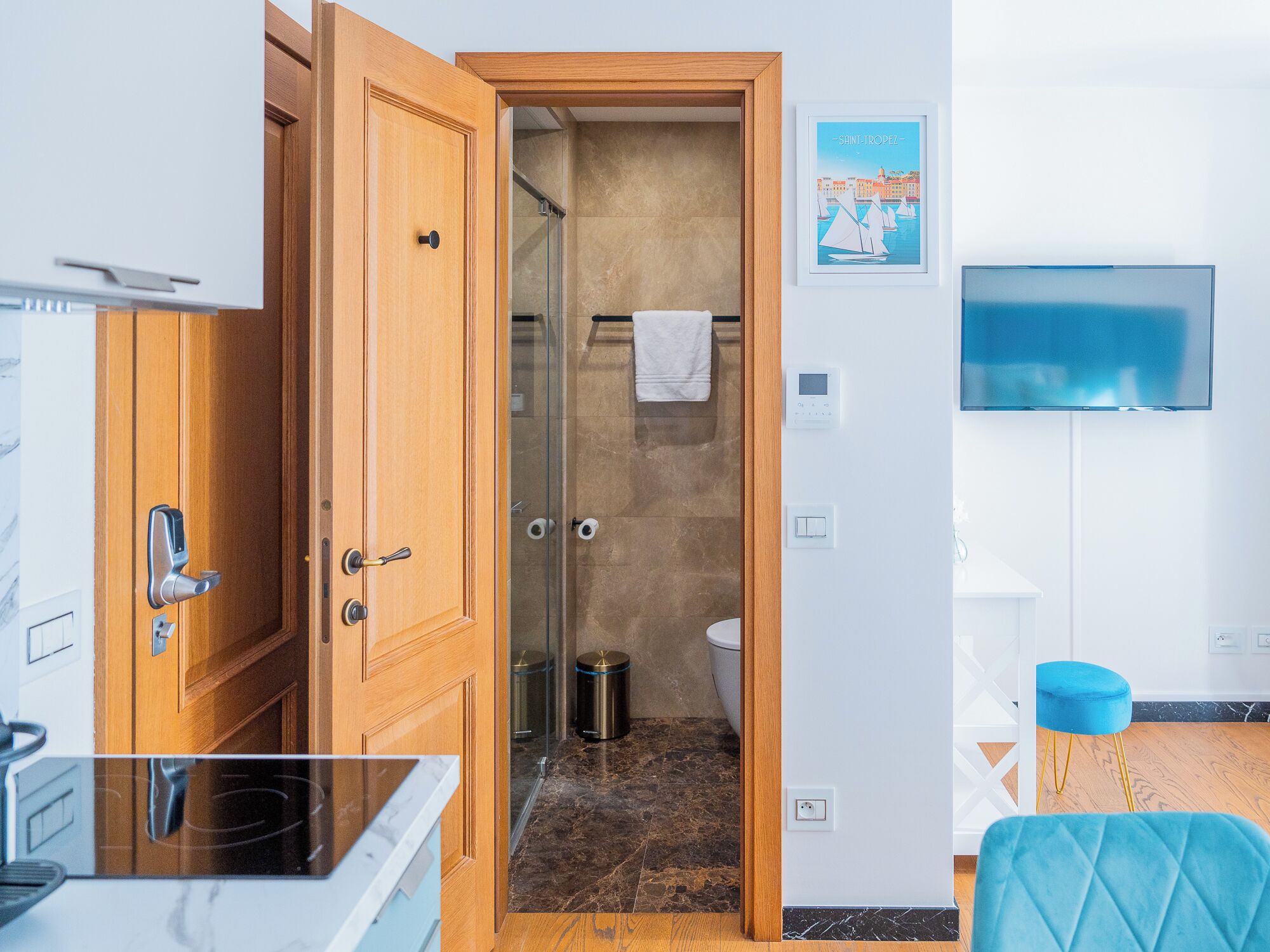 A hotel room with a wooden door, a shower, a toilet, a TV, and a blue chair.