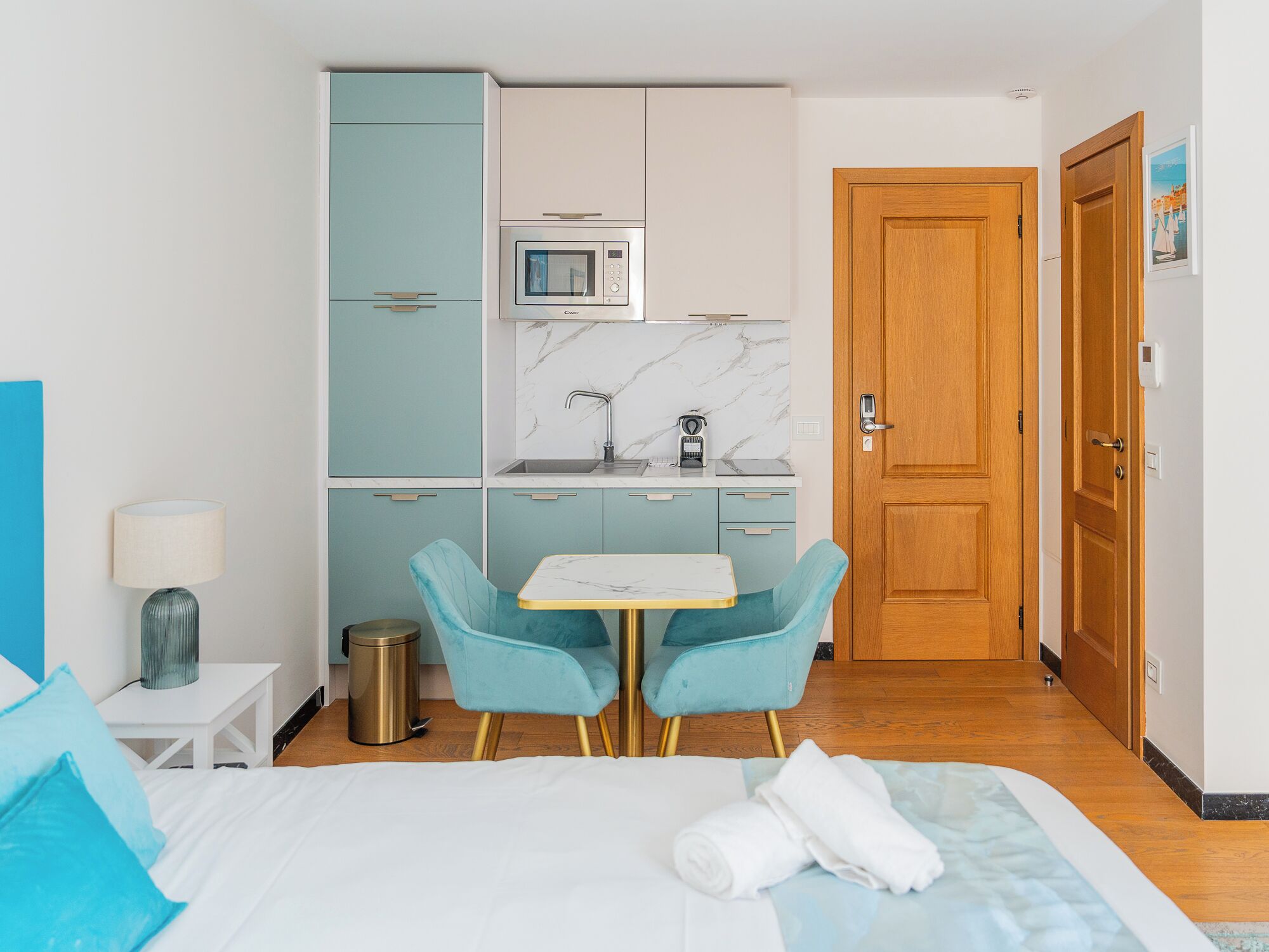 Una camera d'albergo compatta con un letto, un piccolo tavolo con sedie, un angolo cottura e una porta in legno.