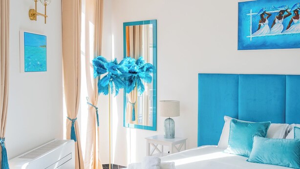 Una habitación de hotel moderna con una cama grande, cabecero azul, un cuadro, un espejo y una ventana con cortinas.