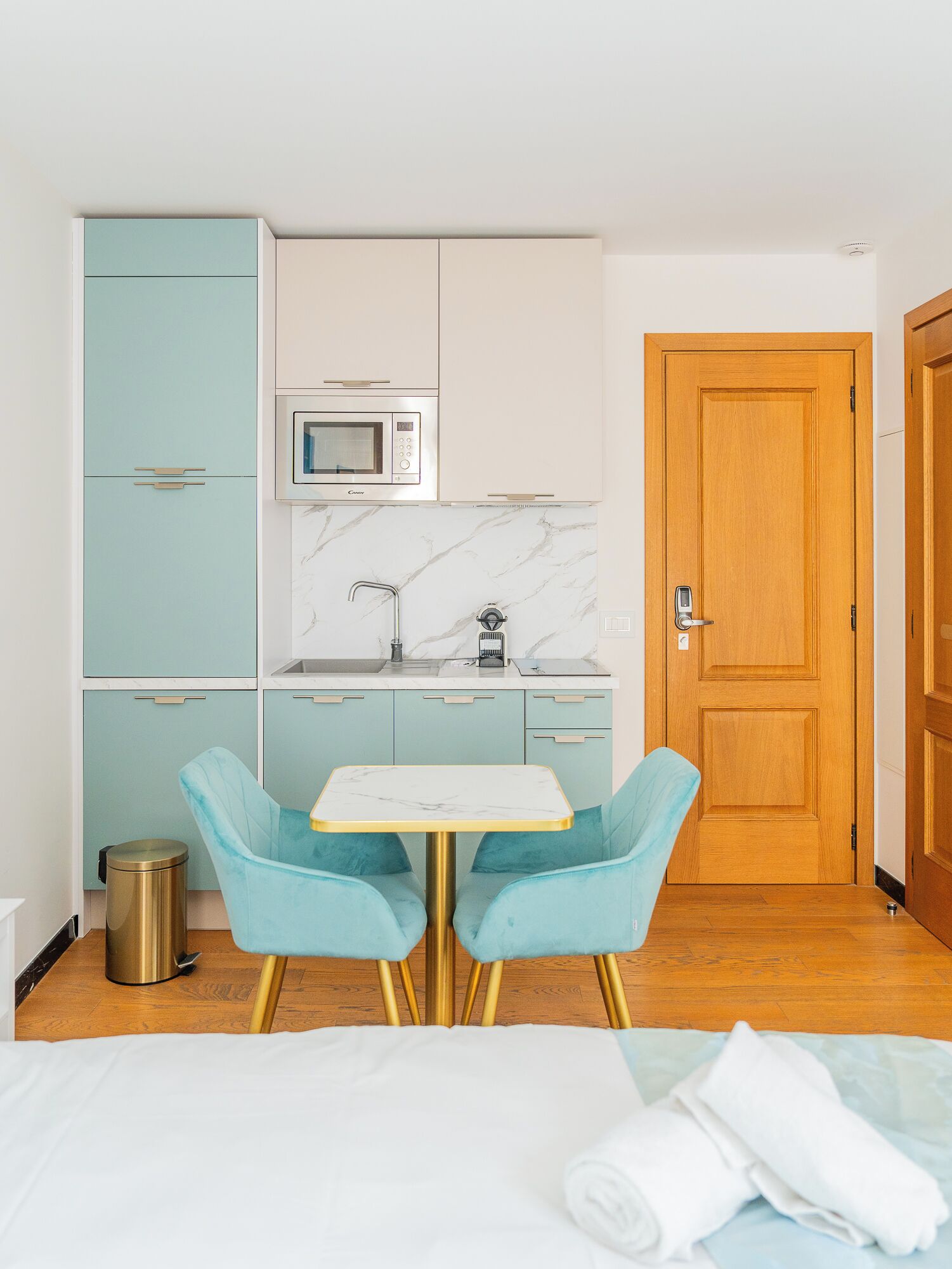 Cocina compacta con microondas, fregadero y armarios. Se observa una pequeña mesa con dos sillas y una cama con manta blanca.