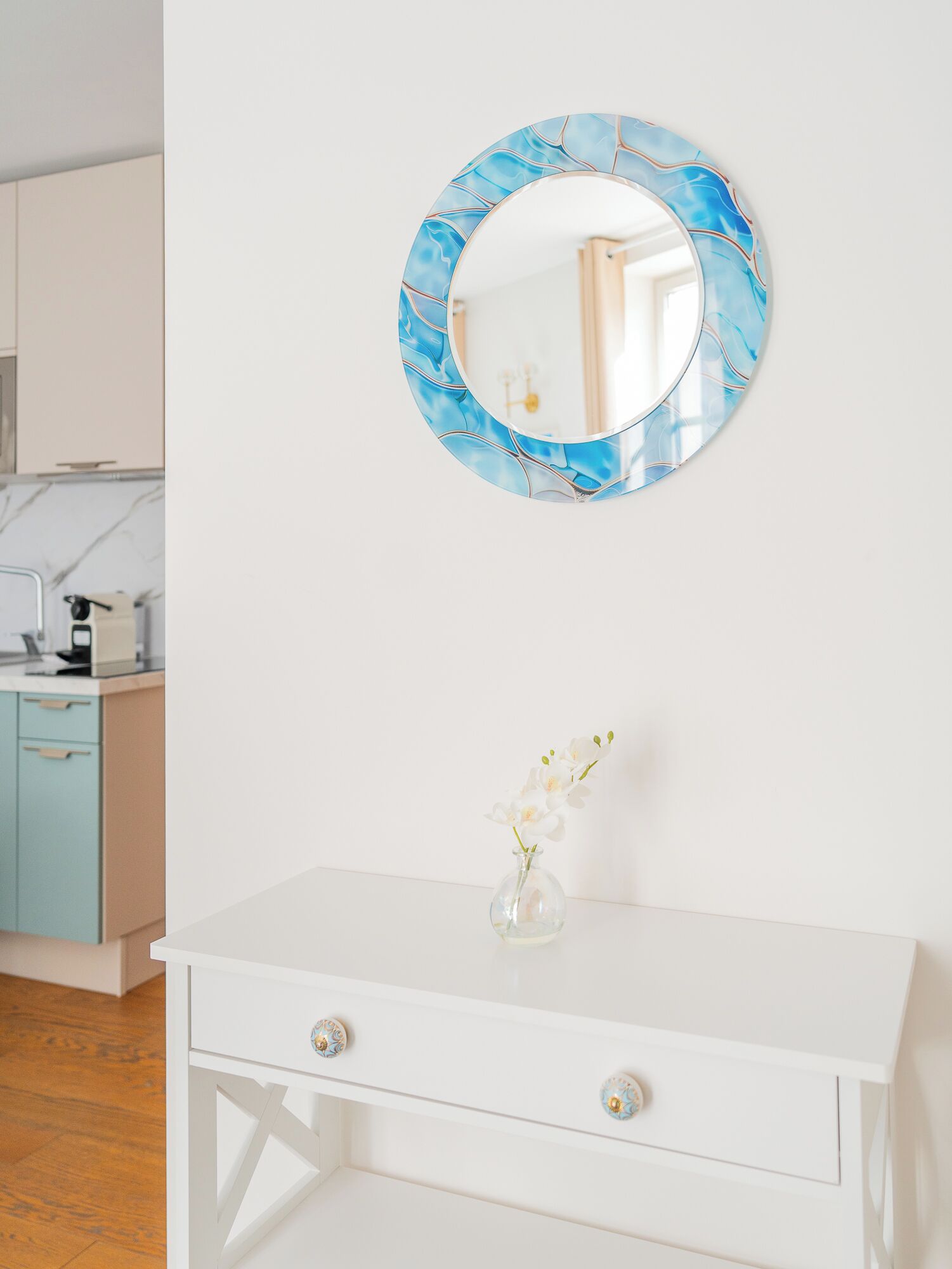 A white dresser with a vase of white flowers, a round mirror with a blue marble pattern, and a kitchen in the background.