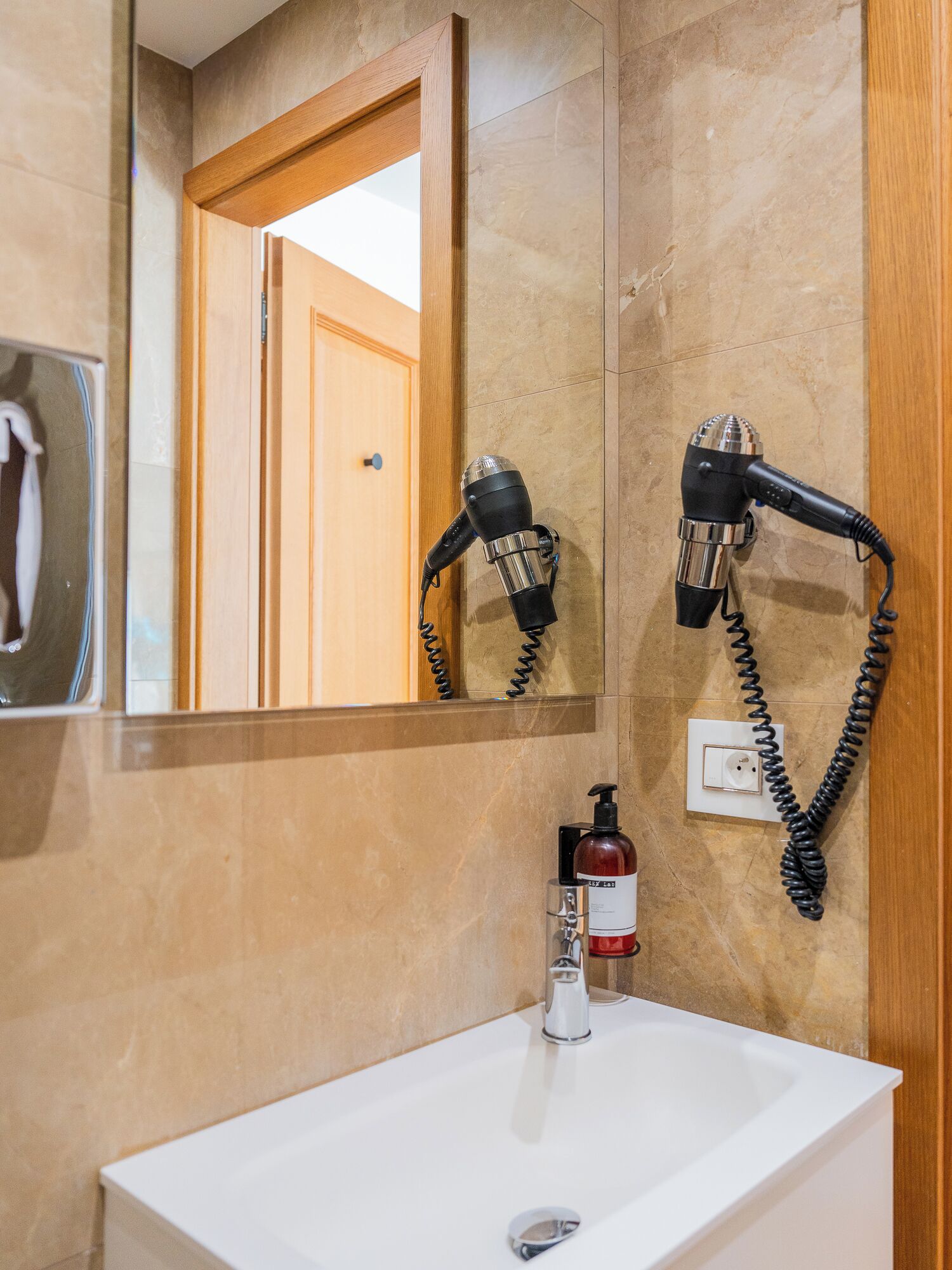 A bathroom with a white sink, a mirror, two wall-mounted hair dryers, a bottle of hand soap, and a towel rack.