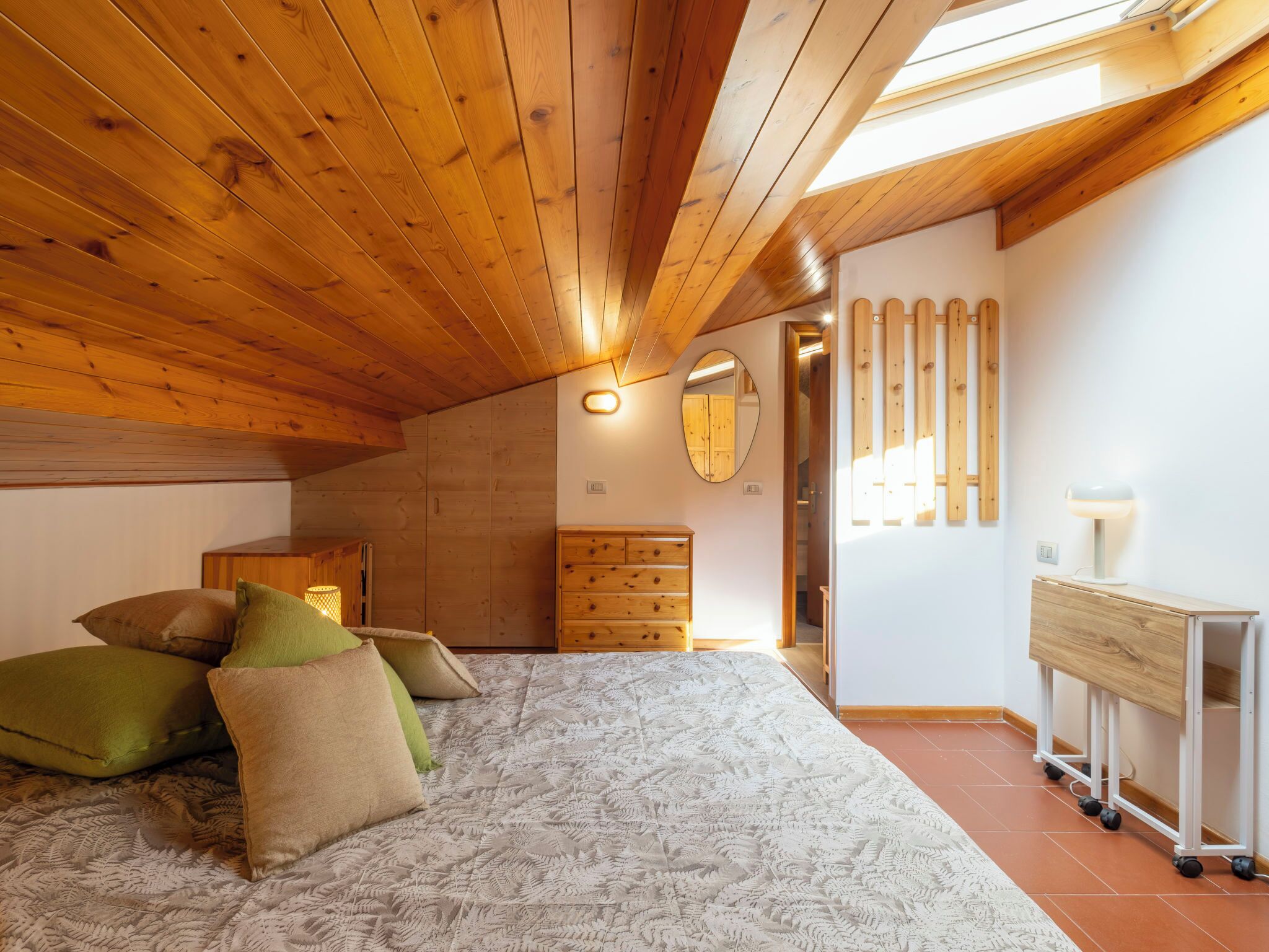 Ein Schlafzimmer mit schrägen Wänden, Holzwänden, einem Bett mit gemusterten Bettbezügen, einem Schminktisch, einem Spiegel und einem Schreibtisch mit Lampe.
