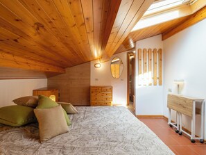 Ein Schlafzimmer mit schrägen Wänden, Holzwänden, einem Bett mit gemusterten Bettbezügen, einem Schminktisch, einem Spiegel und einem Schreibtisch mit Lampe.