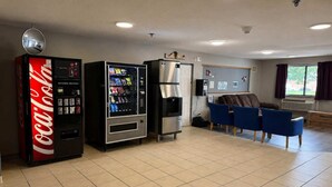 A room with a Coca-Cola vending machine, a refrigerator, blue chairs, and a window with curtains.