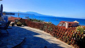 Eine terrasse mit Steinplatten und Blick auf das Meer, ein Holzzaun und ein Gebäude mit rotem Dach.