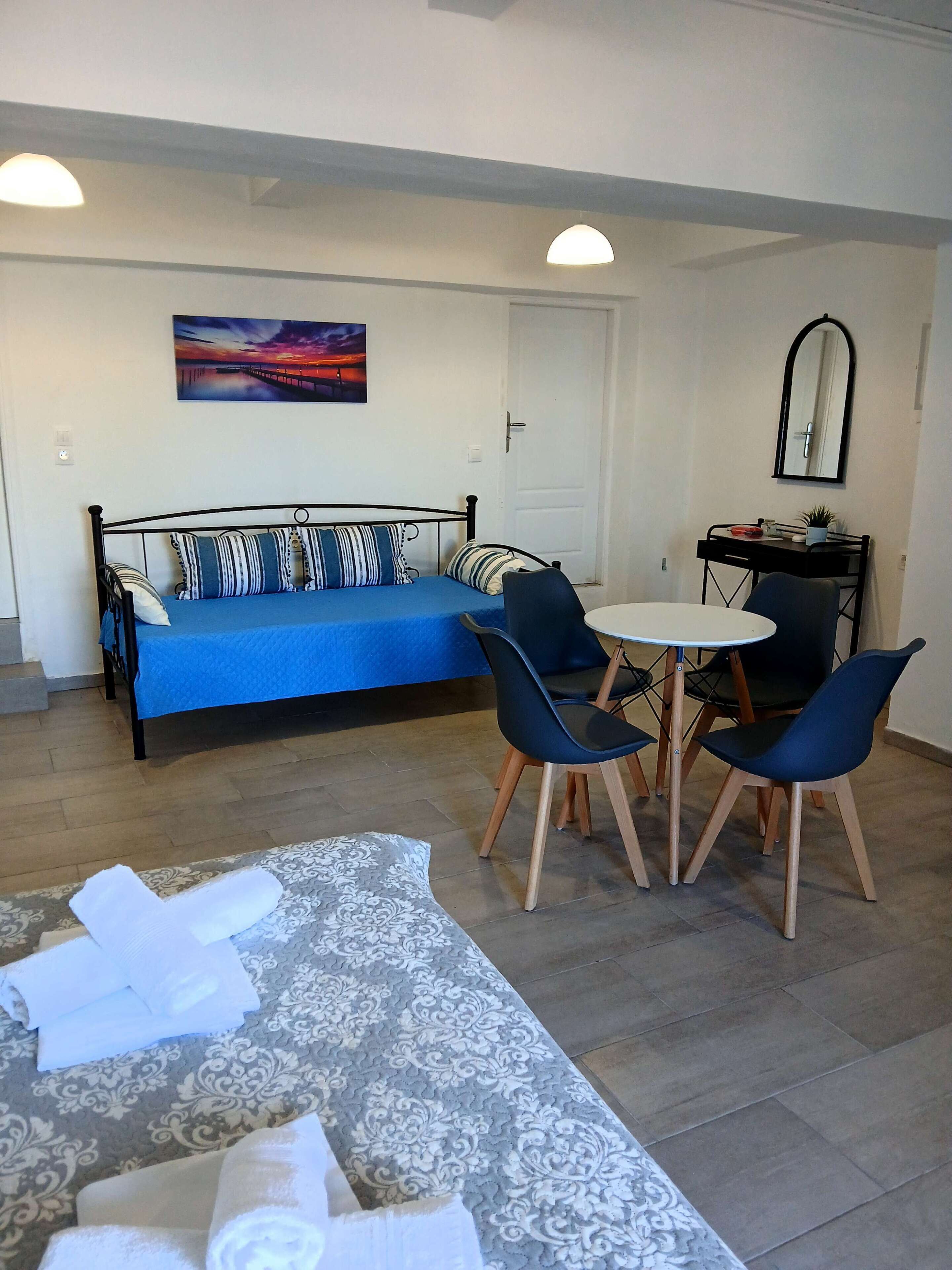 A room with a bed, a sofa, a dining table with chairs, a small kitchen area, and a painting on the wall.