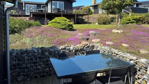 Un patio con mesa de vidrio y sillas de mimbre, rodeado por un muro de piedra y un jardín con flores moradas.