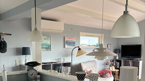 Cocina moderna con una isla central, luces colgantes y vistas a un salón con televisor y asientos.