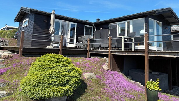 Una casa moderna con terraza, mobiliario exterior y jardín con flores púrpuras.