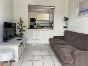 A living room with a flat-screen TV, a sofa, a small white stool, a potted plant, and a kitchen visible through an open door. - The Alizées Getaway (Le Château-d'Oléron)