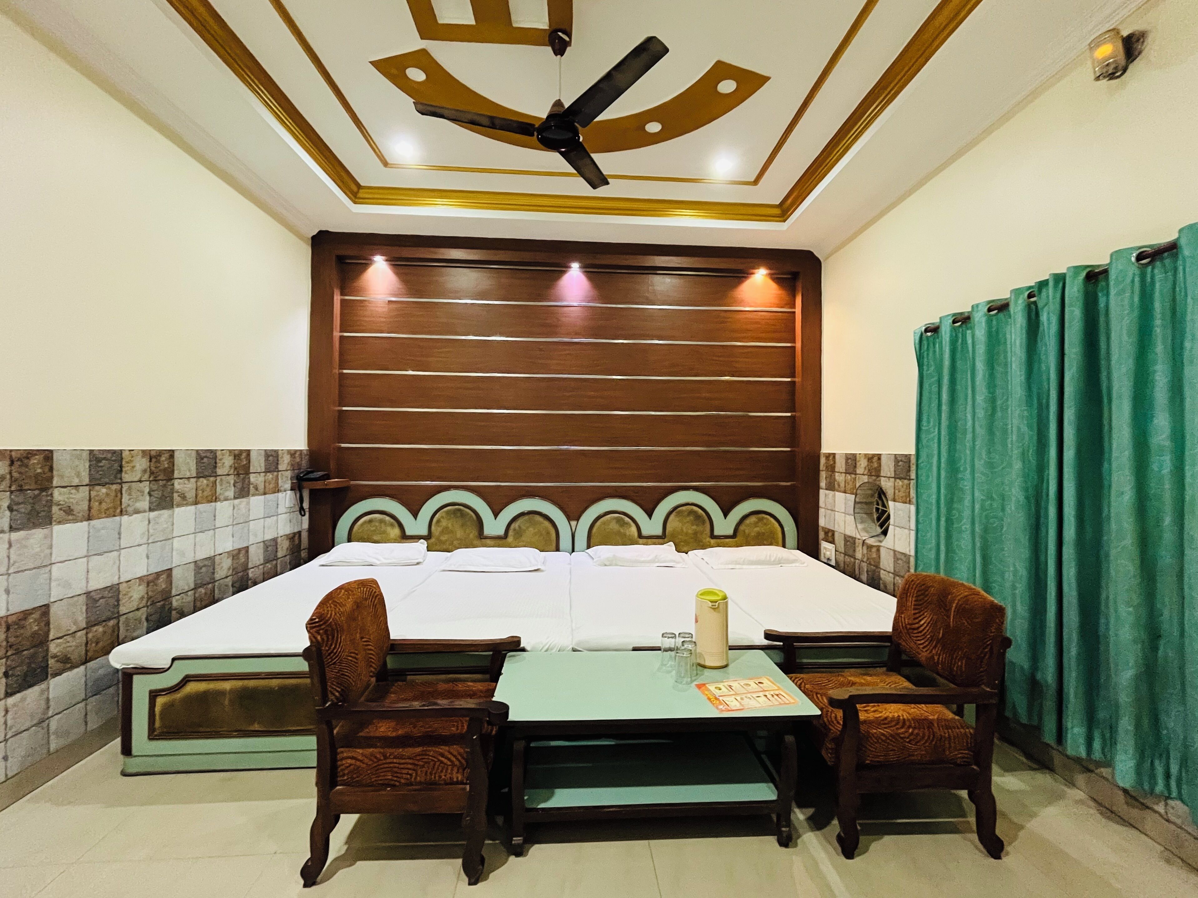 Une chambre d’hôtel avec un grand lit, deux chaises, une petite table, un ventilateur de plafond et un rideau vert.