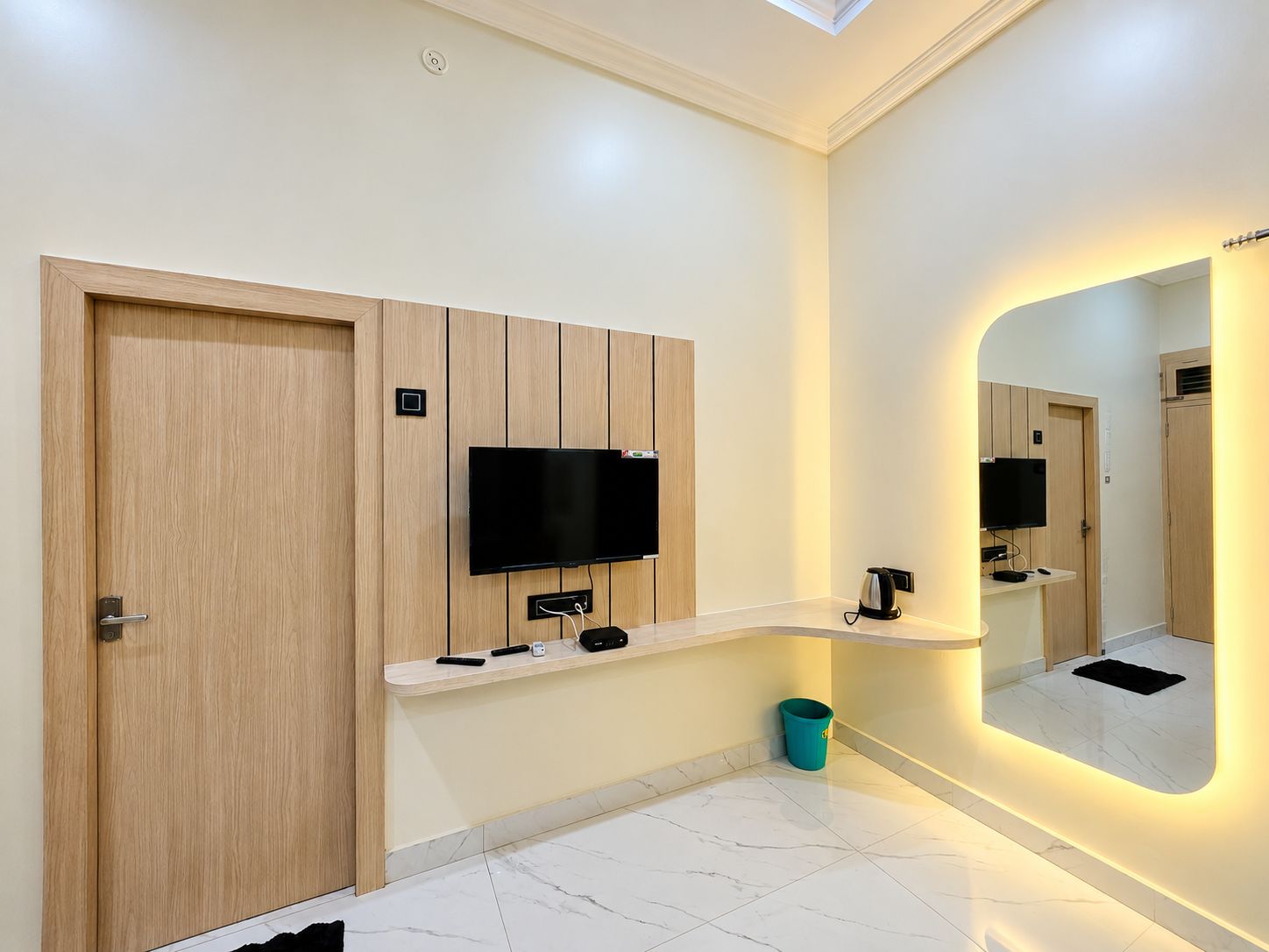 A modern bathroom with a wooden door, a mounted TV, a mirror with integrated lighting, and a marble floor.