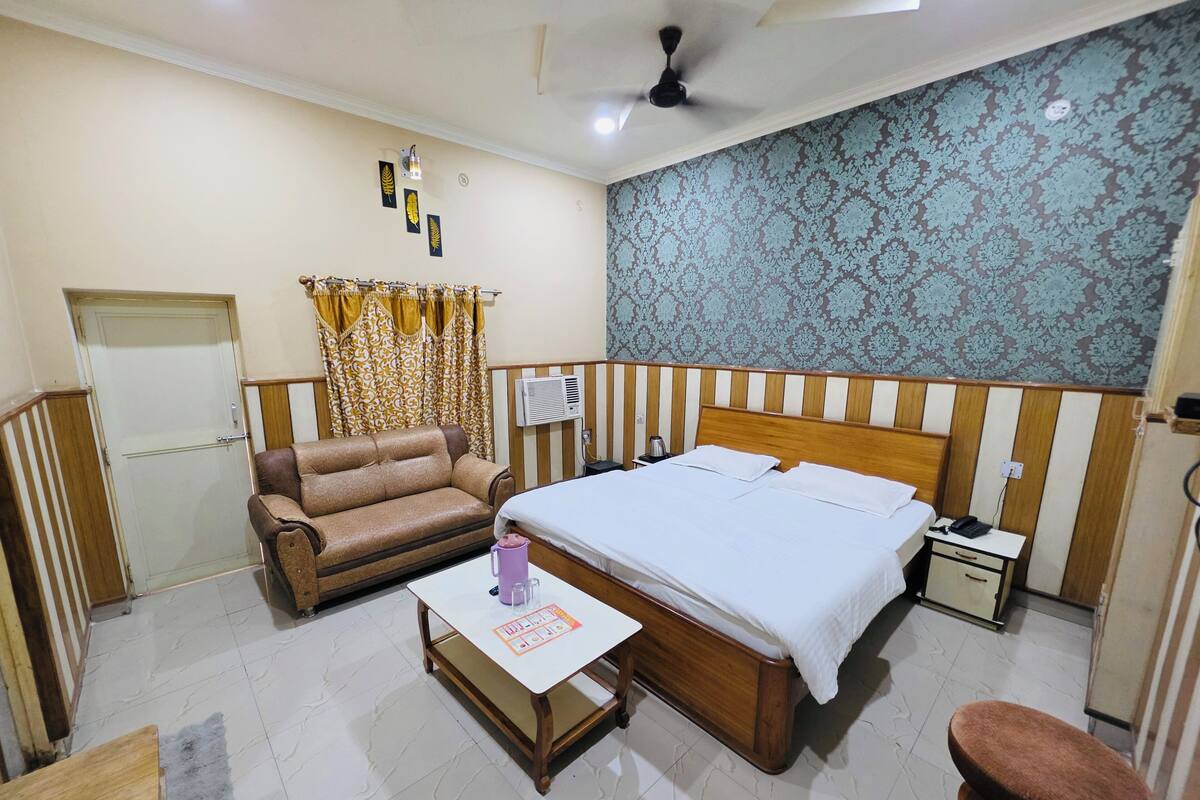 A bedroom with a bed, a sofa, a table, a chair, curtains, a ceiling fan, and a patterned wall.