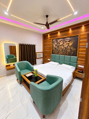 Habitación de hotel moderna con una cama, dos sillones verdes, una mesita con una lámpara, un ventilador de techo y una obra de arte en la pared con elefantes.