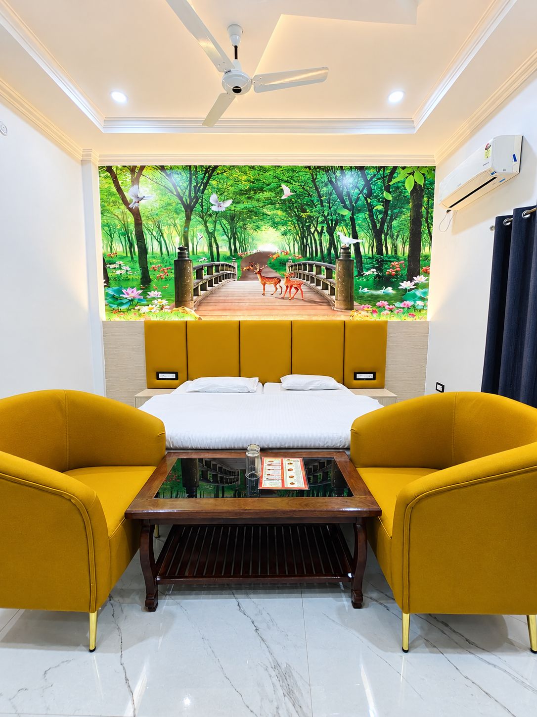 A room with a bed, two yellow armchairs, a coffee table, a ceiling fan, and a wall mural of a forest scene.