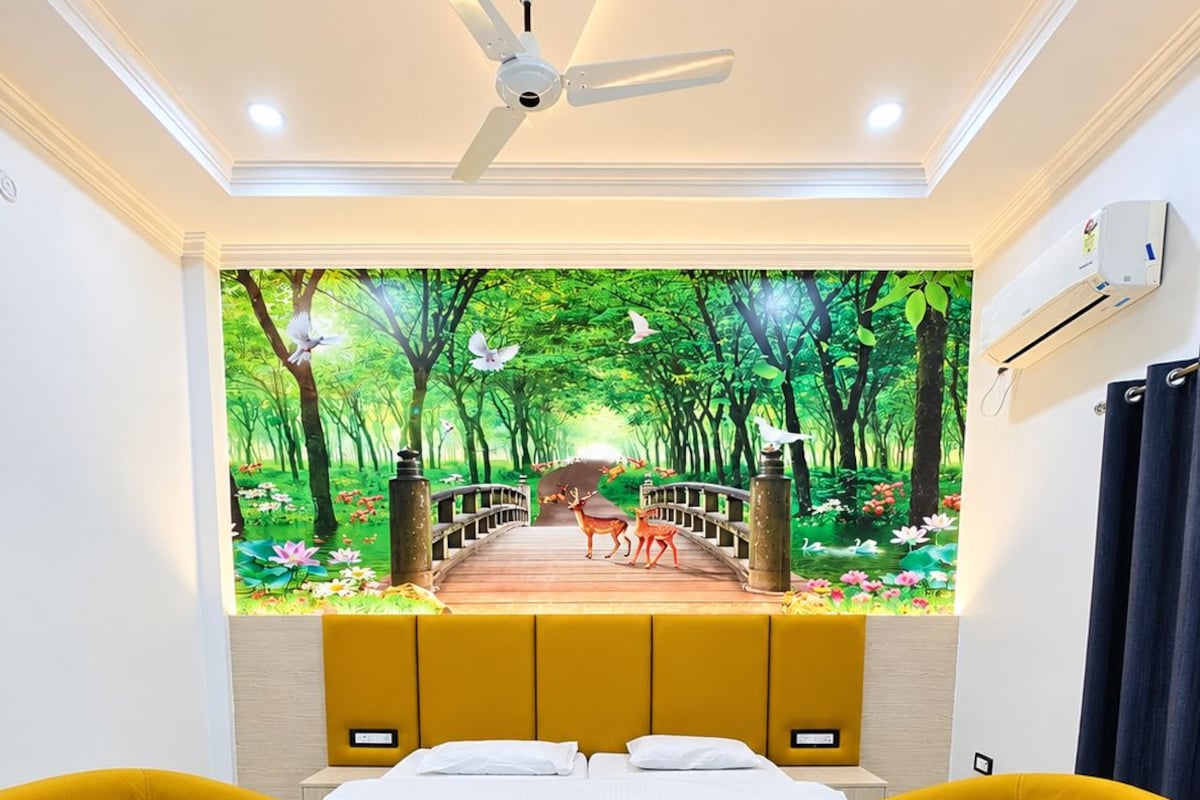 Una habitación con una cama, dos sillones amarillos, una mesa de centro, un ventilador de techo y un mural en la pared con una escena de bosque.