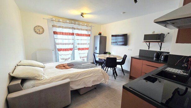 Ein kleines Hotelzimmer mit Bett, Esstisch mit Stühlen, einer Küchenzeile, einem Fernseher und einem Fenster mit Vorhängen.